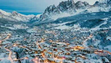 Vista aérea da vila alpina de Cortina d'Ampezzo coberta de neve ao anoitecer, com as montanhas Dolomitas ao fundo e luzes urbanas acesas.