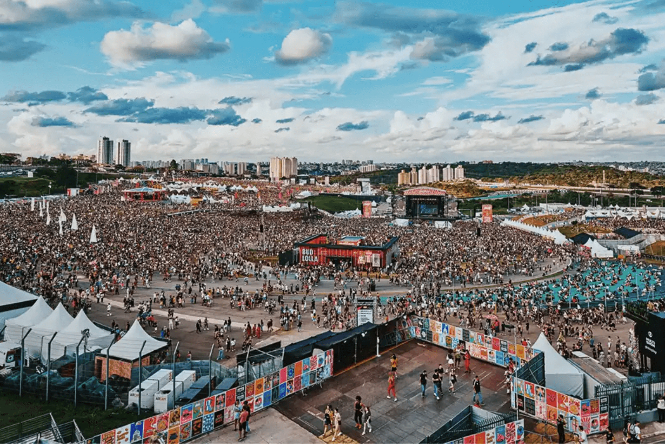 Foto panorâmica aérea de um festival de música em um autódromo. O gramado está coberto por milhares de pessoas entre tendas brancas, palcos e estruturas de ativação de marcas.