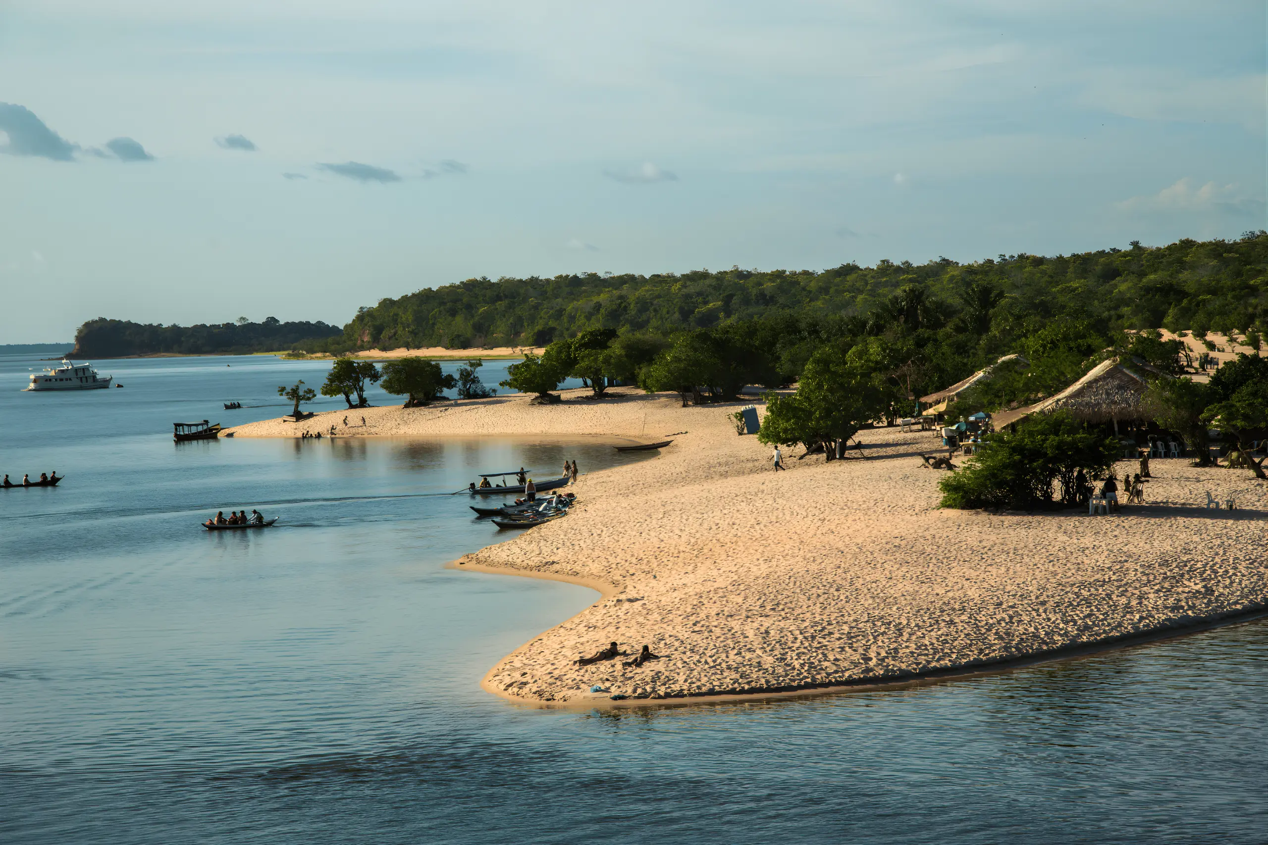 Fotografia aérea de uma ponta de areia clara avançando sobre o Rio Tapajós, com barcos ancorados e quiosques de palha sob árvores.