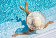 Mulher com chapéu de palha sentada à beira de piscina azul em dia ensolarado vista de cima