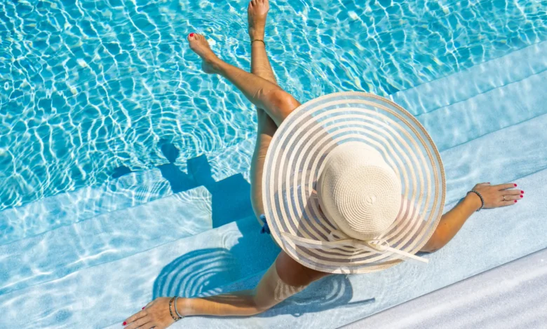 Mulher com chapéu de palha sentada à beira de piscina azul em dia ensolarado vista de cima