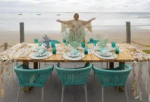 Martha Medeiros de braços abertos atrás de mesa decorada com elementos marinhos na praia de Milagres em Alagoas