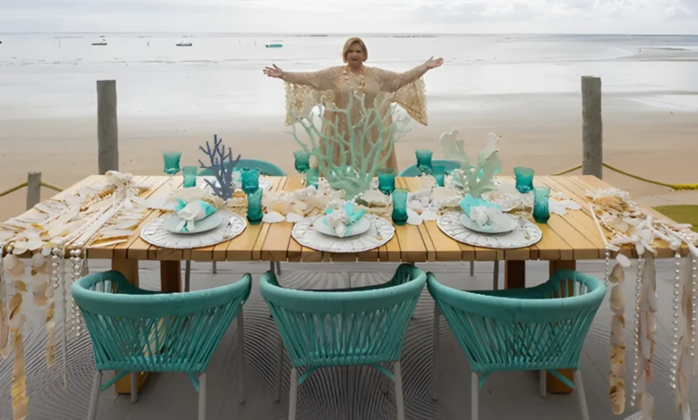 Martha Medeiros de braços abertos atrás de mesa decorada com elementos marinhos na praia de Milagres em Alagoas