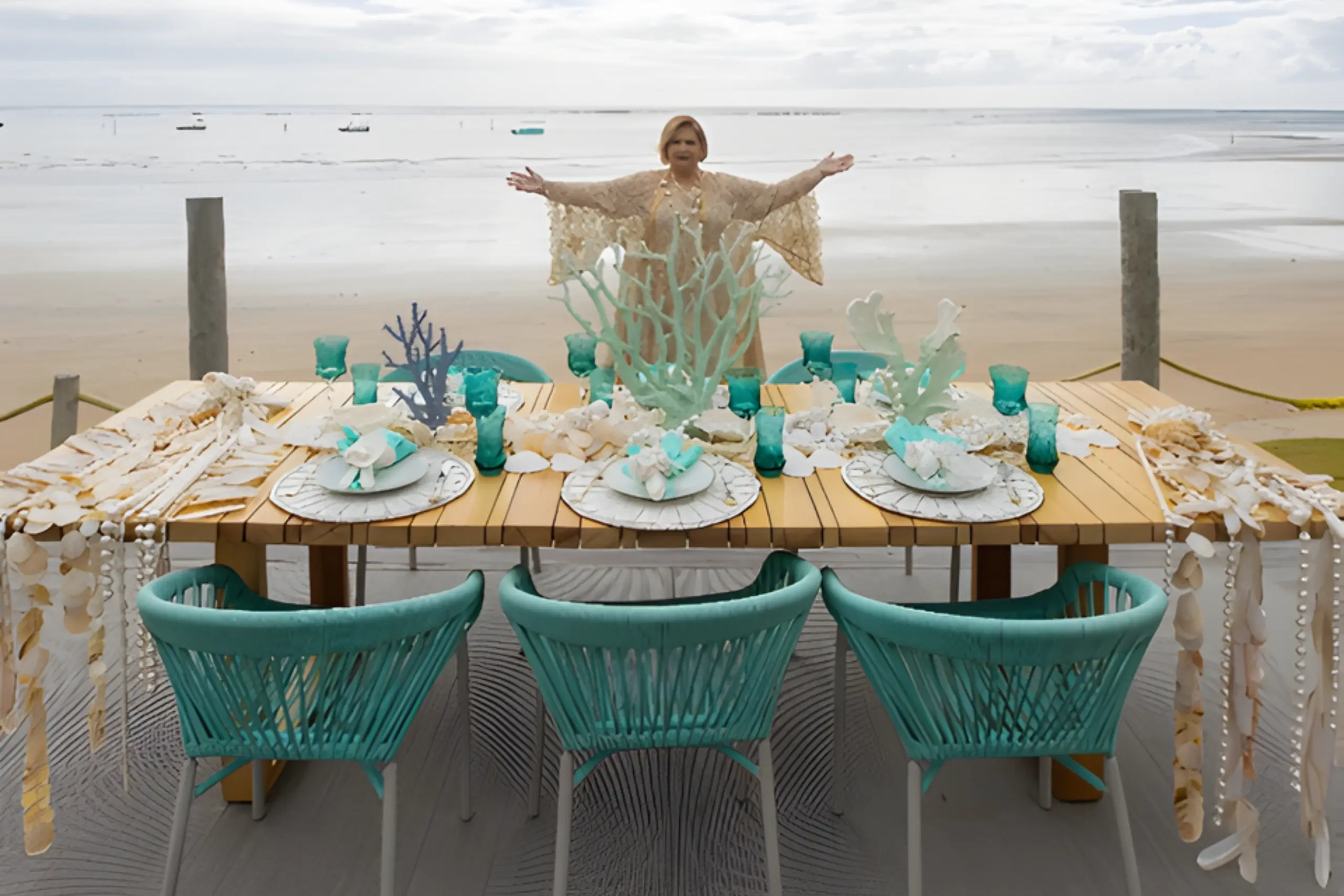 Martha Medeiros de braços abertos atrás de mesa decorada com elementos marinhos na praia de Milagres em Alagoas