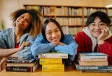 Três jovens estudantes internacionais sorrindo apoiados em pilhas de livros dentro de uma biblioteca