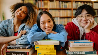 Três jovens estudantes internacionais sorrindo apoiados em pilhas de livros dentro de uma biblioteca