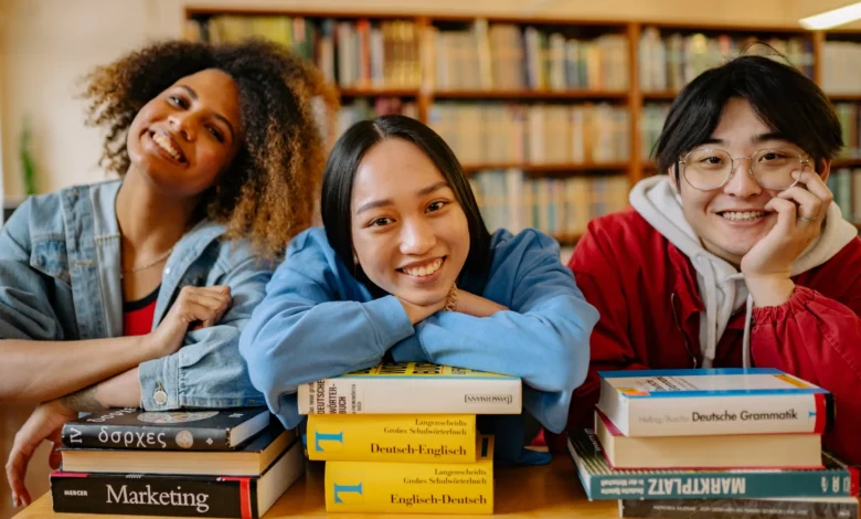 Três jovens estudantes internacionais sorrindo apoiados em pilhas de livros dentro de uma biblioteca