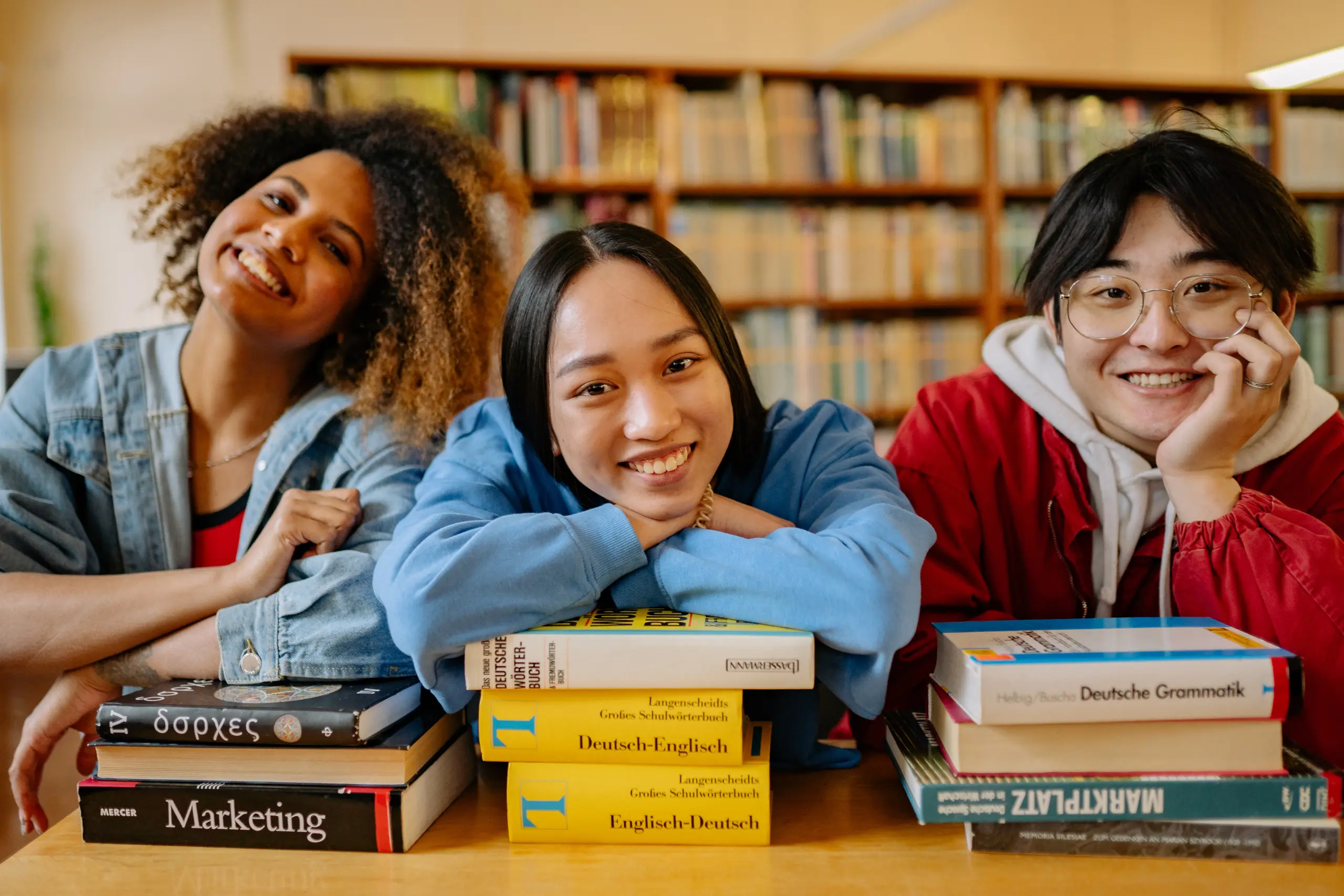 Três jovens estudantes internacionais sorrindo apoiados em pilhas de livros dentro de uma biblioteca