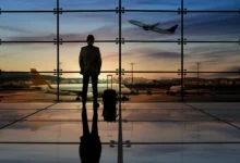 Silhueta de executivo com mala de viagem em frente à janela de aeroporto com aviões e céu alaranjado ao fundo