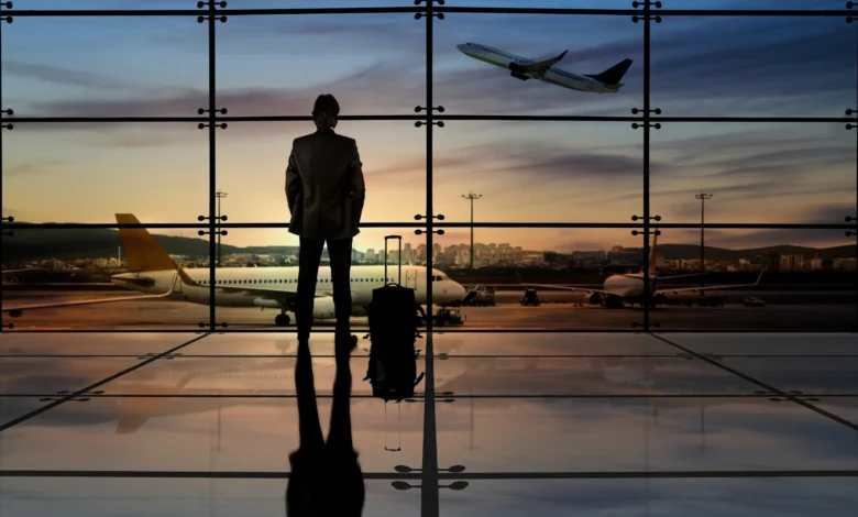 Silhueta de executivo com mala de viagem em frente à janela de aeroporto com aviões e céu alaranjado ao fundo