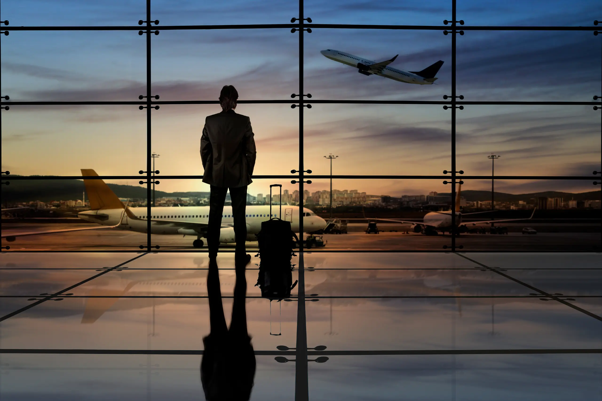 Silhueta de executivo com mala de viagem em frente à janela de aeroporto com aviões e céu alaranjado ao fundo