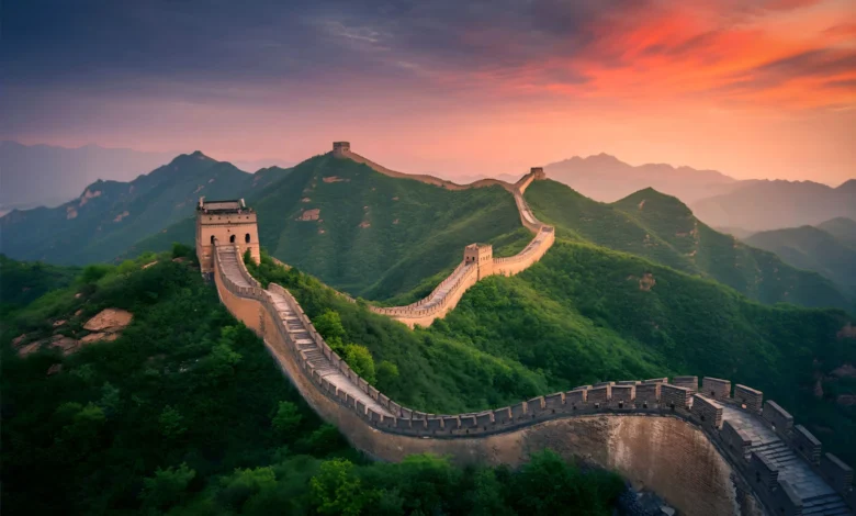 Vista aérea da Grande Muralha da China serpenteando por montanhas verdes ao entardecer com céu em tons de laranja e roxo