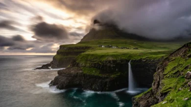 Falésia verde com cascata despencando no mar nas Ilhas Faroé sob céu dramático com nuvens escuras e aldeia ao fundo