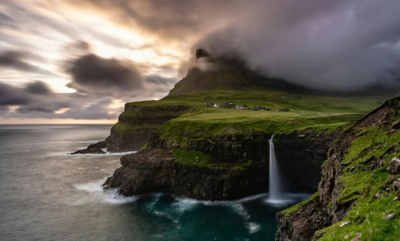 Falésia verde com cascata despencando no mar nas Ilhas Faroé sob céu dramático com nuvens escuras e aldeia ao fundo
