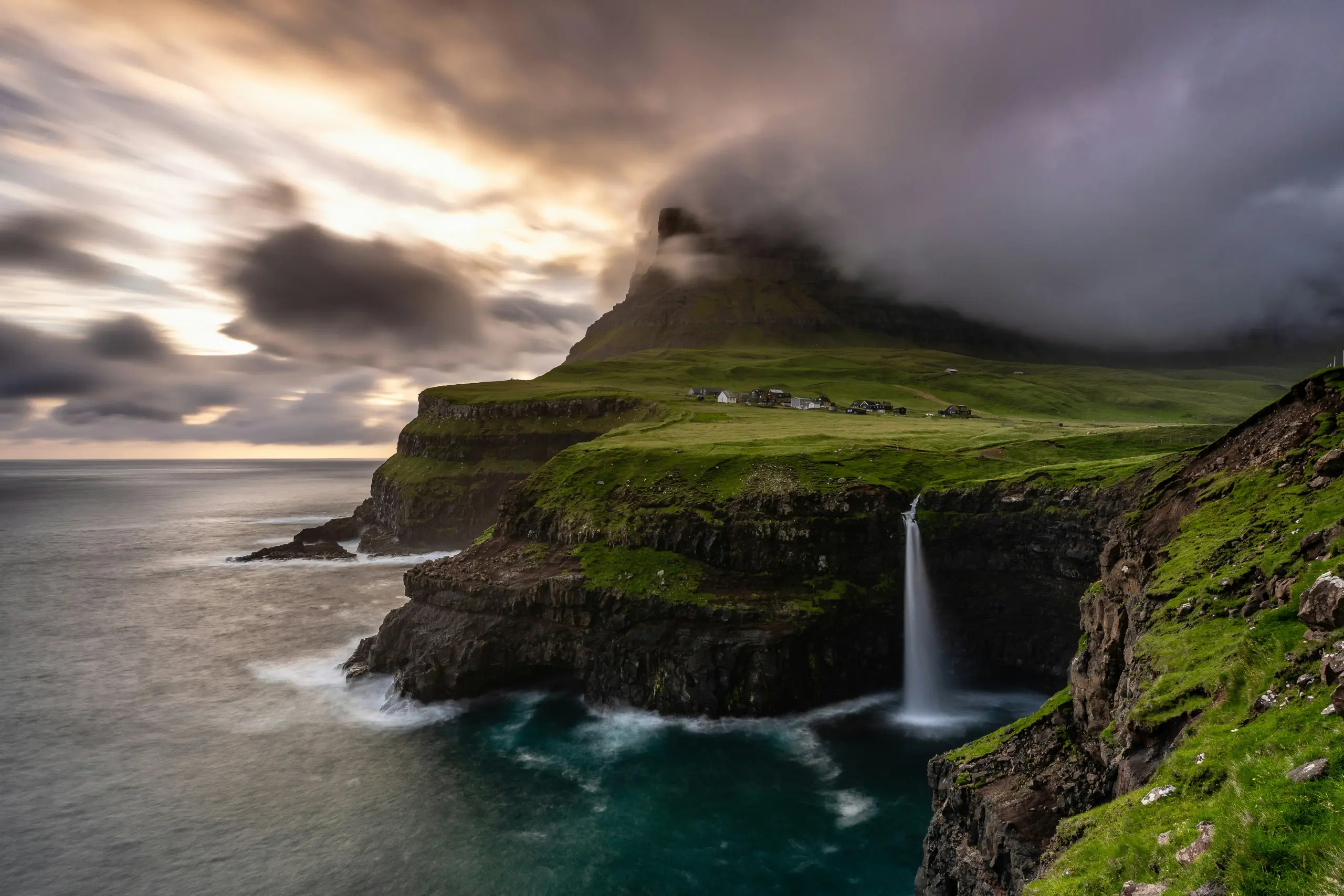 Falésia verde com cascata despencando no mar nas Ilhas Faroé sob céu dramático com nuvens escuras e aldeia ao fundo
