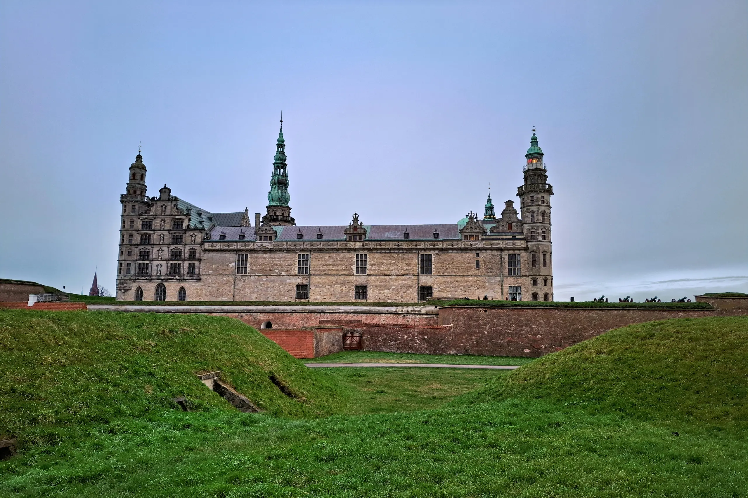 Vista frontal do Kronborg Castle em Helsingør na Dinamarca com muralhas de grama verde e torres com cúpulas verdes sob céu nublado
