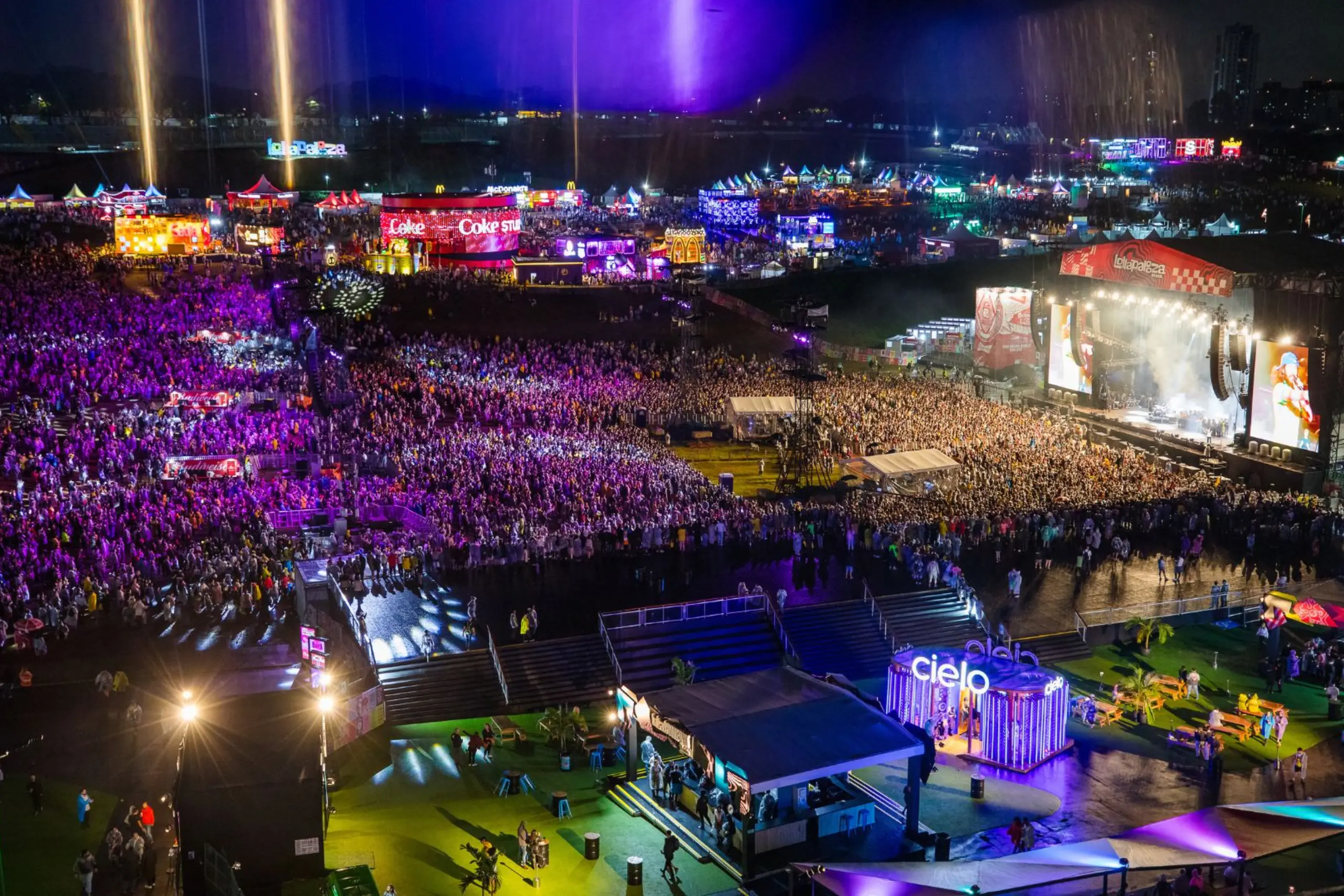 Vista aérea noturna do Lollapalooza Brasil no Autódromo de Interlagos com multidão e palco iluminado em tons de roxo e laranja