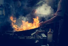 Chef trabalhando sob pressão em cozinha profissional com chamas altas em wok e fumaça no ambiente escuro