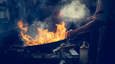 Chef trabalhando sob pressão em cozinha profissional com chamas altas em wok e fumaça no ambiente escuro