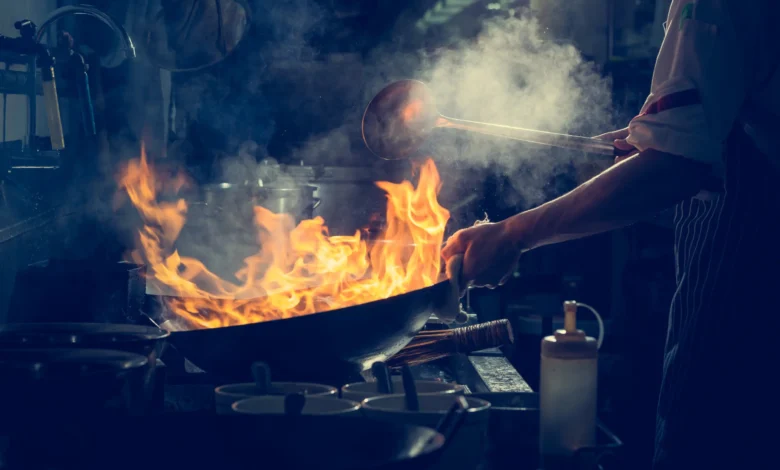 Chef trabalhando sob pressão em cozinha profissional com chamas altas em wok e fumaça no ambiente escuro