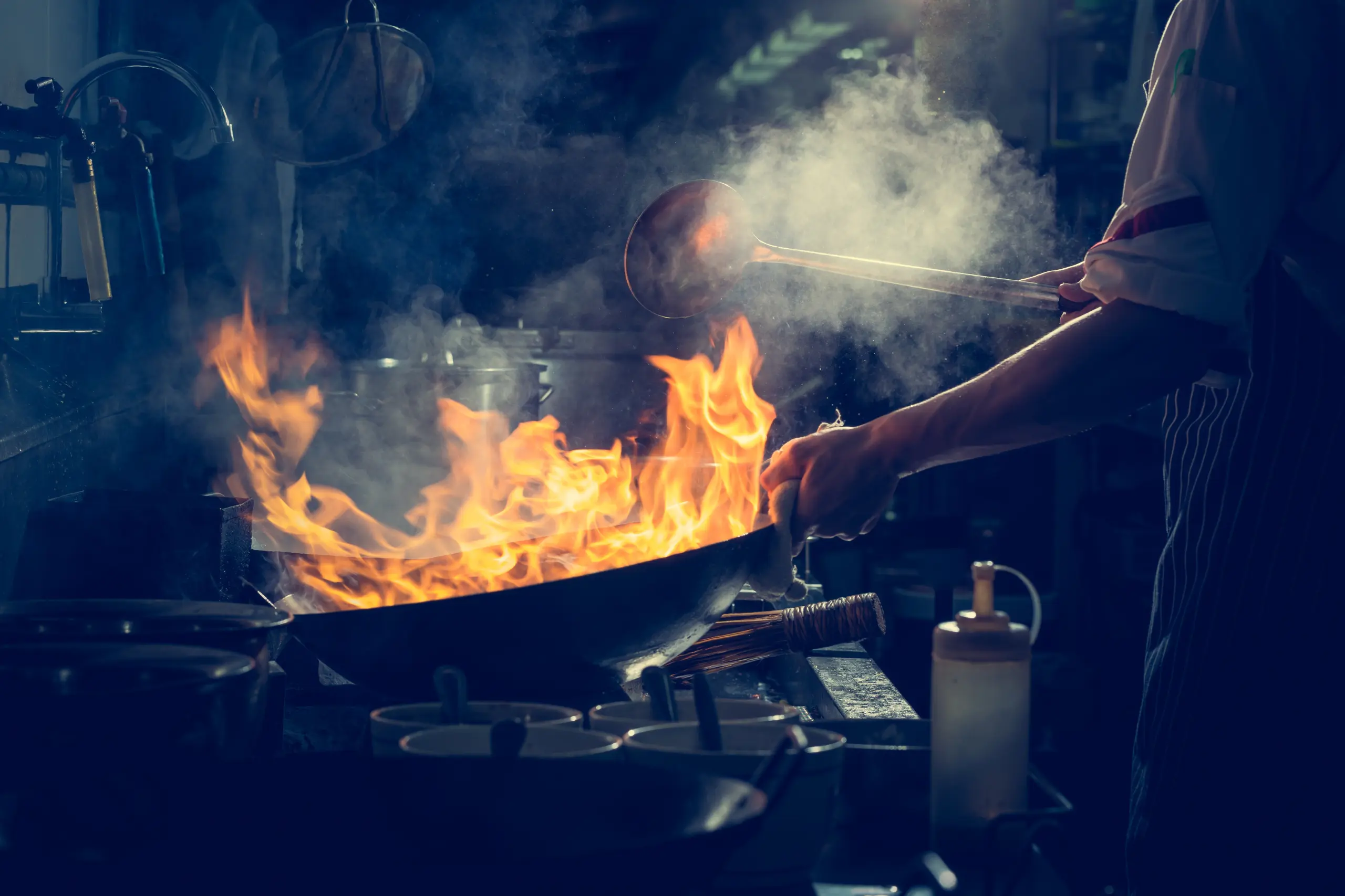 Chef trabalhando sob pressão em cozinha profissional com chamas altas em wok e fumaça no ambiente escuro