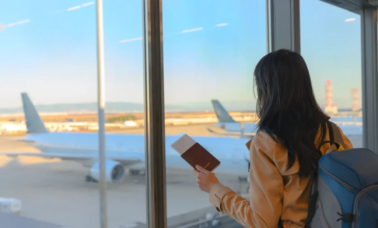 Mulher de costas com mochila azul segurando passaporte em frente à janela do aeroporto com aviões ao fundo
