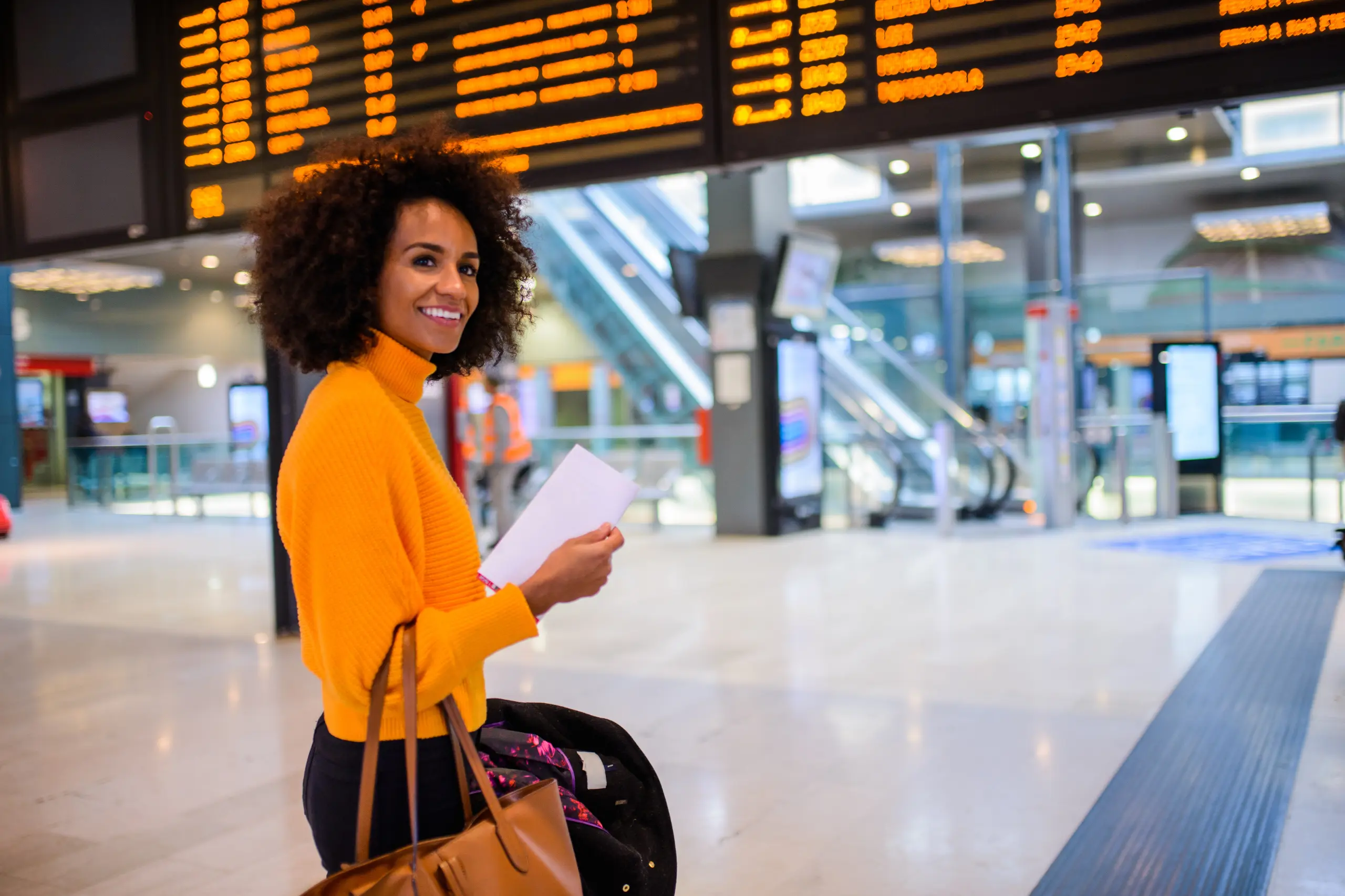 Mulher negra sorrindo com suéter laranja segurando passagem em aeroporto com painel de voos ao fundo