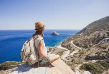 Mulher de costas com mochila e chapéu de palha sentada em pedra contemplando o mar azul e paisagem montanhosa ao fundo