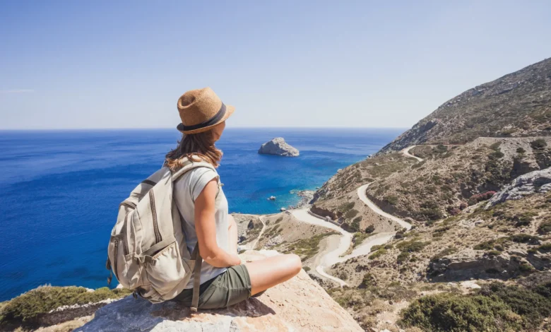 Mulher de costas com mochila e chapéu de palha sentada em pedra contemplando o mar azul e paisagem montanhosa ao fundo