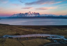 Fotografia aérea do hotel Tierra Patagonia, uma estrutura alongada e curva de madeira que se integra à estepe, com o Lago Sarmiento e as montanhas nevadas de Torres del Paine ao fundo sob um céu de entardecer.