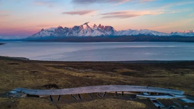 Fotografia aérea do hotel Tierra Patagonia, uma estrutura alongada e curva de madeira que se integra à estepe, com o Lago Sarmiento e as montanhas nevadas de Torres del Paine ao fundo sob um céu de entardecer.