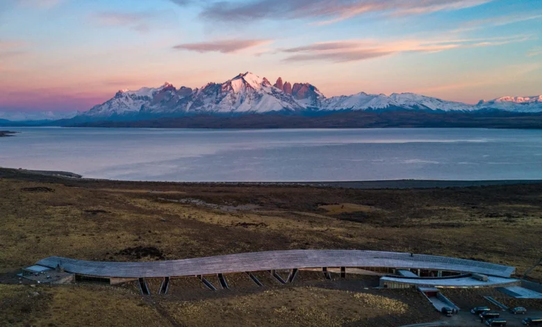 Fotografia aérea do hotel Tierra Patagonia, uma estrutura alongada e curva de madeira que se integra à estepe, com o Lago Sarmiento e as montanhas nevadas de Torres del Paine ao fundo sob um céu de entardecer.