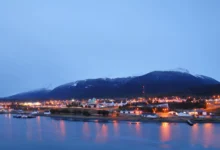 Uma vista noturna de uma cidade costeira aninhada na base de uma grande montanha coberta de neve. A cidade está cheia de luzes douradas e quentes que se estendem ao longo da costa. A montanha ao fundo está envolta em nuvens e neve, com luzes de aeronaves visíveis perto do topo. O primeiro plano é uma extensão de água calma que reflete as luzes da cidade.