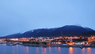Uma vista noturna de uma cidade costeira aninhada na base de uma grande montanha coberta de neve. A cidade está cheia de luzes douradas e quentes que se estendem ao longo da costa. A montanha ao fundo está envolta em nuvens e neve, com luzes de aeronaves visíveis perto do topo. O primeiro plano é uma extensão de água calma que reflete as luzes da cidade.