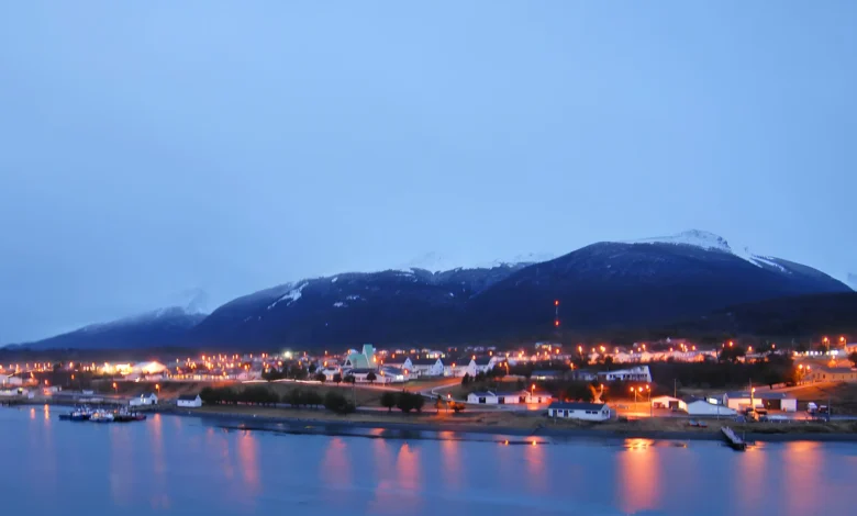 Uma vista noturna de uma cidade costeira aninhada na base de uma grande montanha coberta de neve. A cidade está cheia de luzes douradas e quentes que se estendem ao longo da costa. A montanha ao fundo está envolta em nuvens e neve, com luzes de aeronaves visíveis perto do topo. O primeiro plano é uma extensão de água calma que reflete as luzes da cidade.