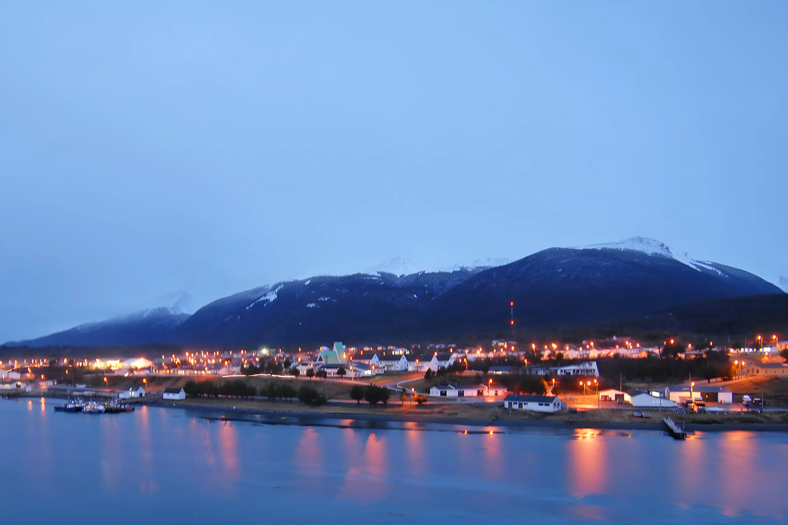 Uma vista noturna de uma cidade costeira aninhada na base de uma grande montanha coberta de neve. A cidade está cheia de luzes douradas e quentes que se estendem ao longo da costa. A montanha ao fundo está envolta em nuvens e neve, com luzes de aeronaves visíveis perto do topo. O primeiro plano é uma extensão de água calma que reflete as luzes da cidade.