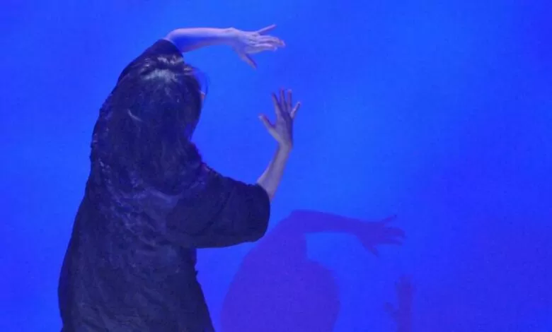 A performer Emilie Sugai está em cena de costas para a câmera, veste um figurino preto, tendo ao fundo um cenário na cor azul.