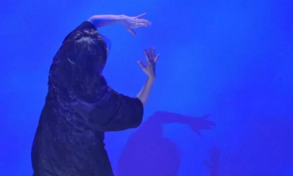 A performer Emilie Sugai está em cena de costas para a câmera, veste um figurino preto, tendo ao fundo um cenário na cor azul.